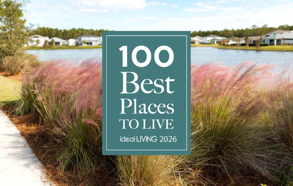 A sign reading "100 Best Places to Live ideaLIVING 2026" stands in front of a path, ornamental grass, a pond, and houses in a suburban neighborhood.