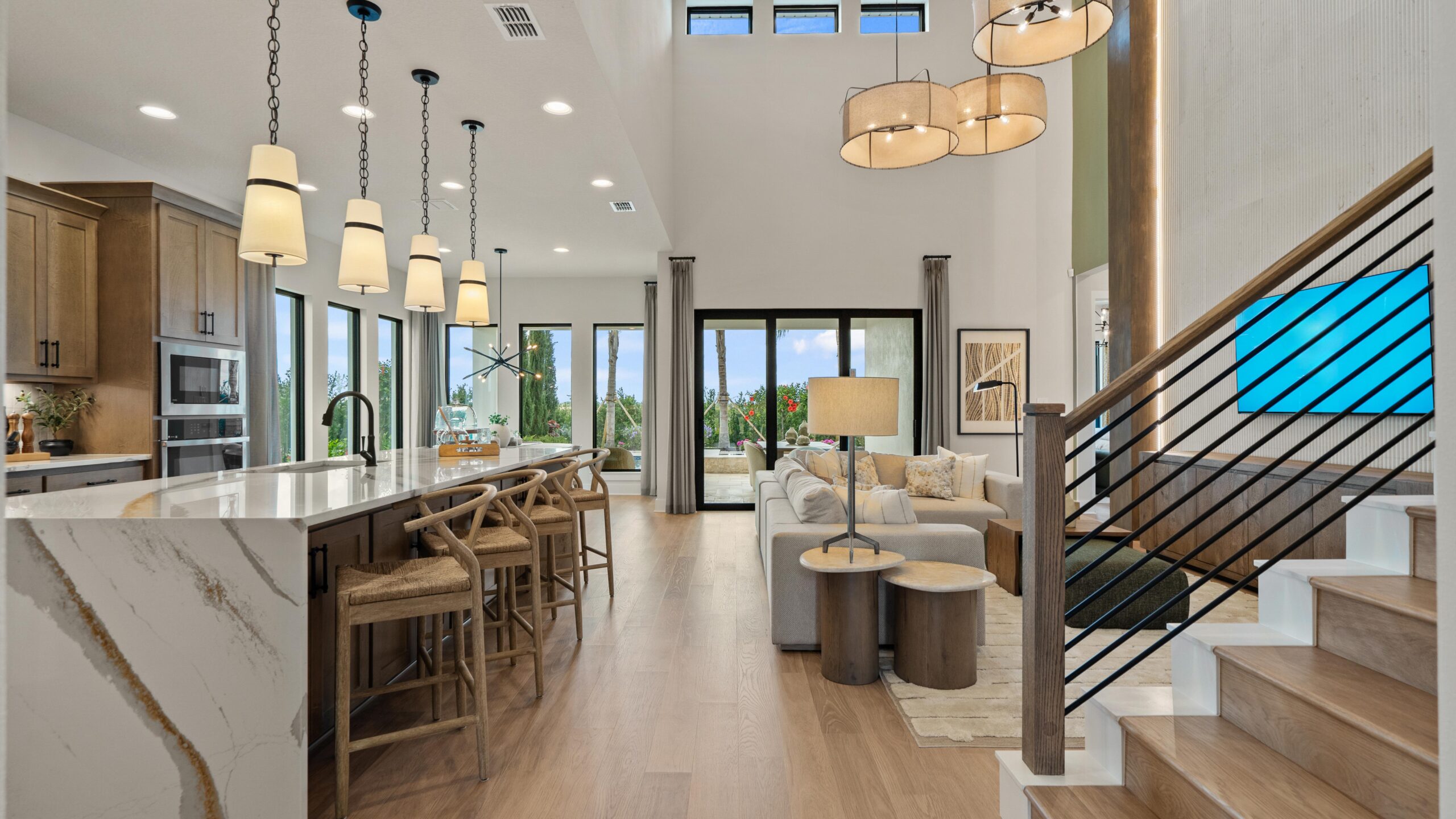Modern open-concept living area with kitchen, island seating, pendant lights, wood floors, and large windows leading to an outdoor space. Stairs with a black railing are visible on the right.